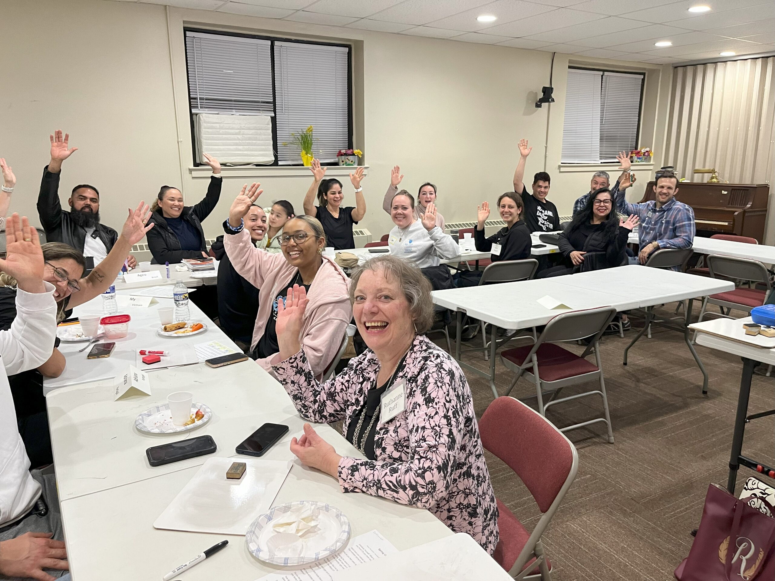A class of students and teachers sitting down at tables and waving to the camera.
