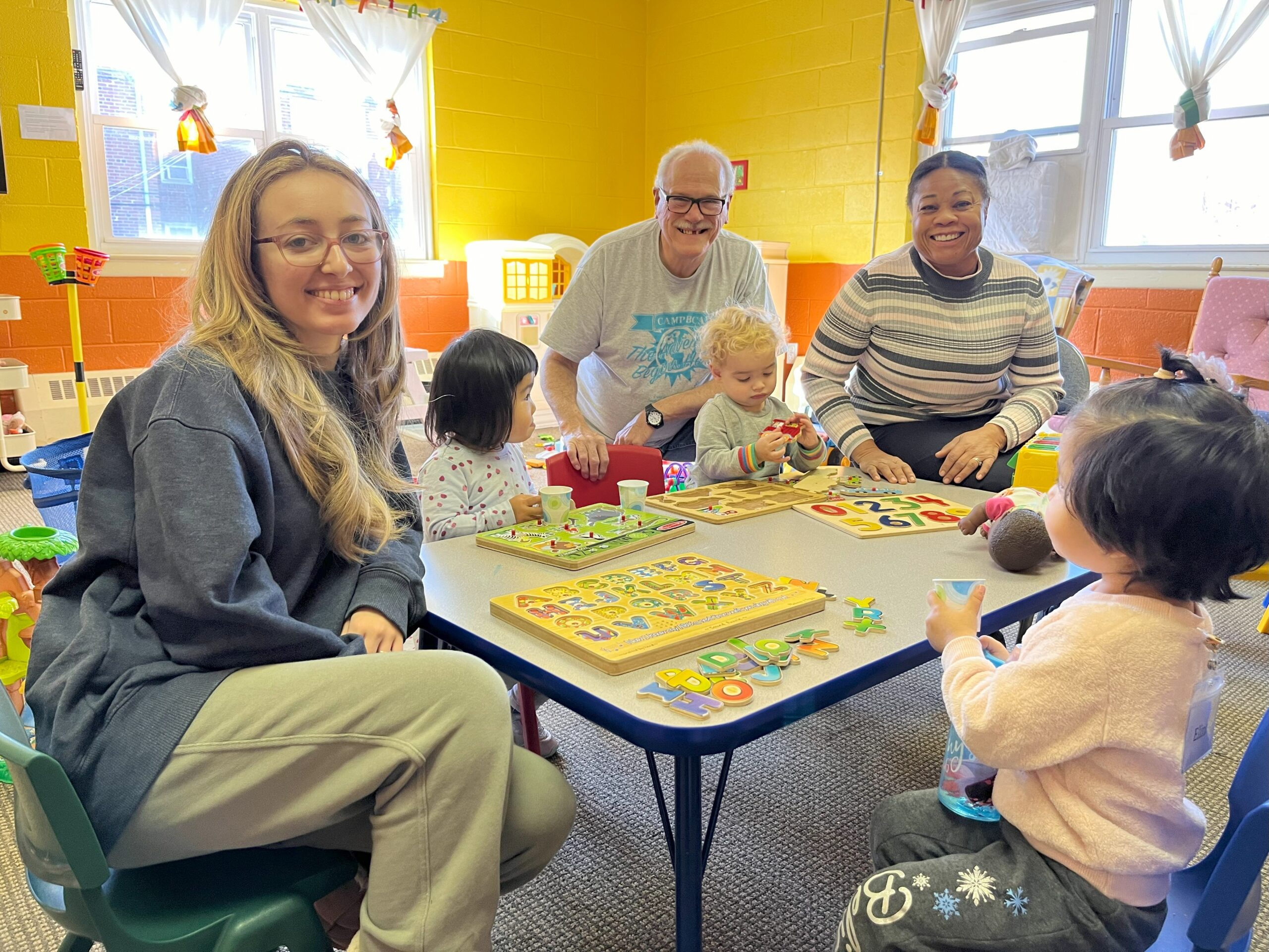 A group of volunteer adults sitting at a table with young children in the nursery.