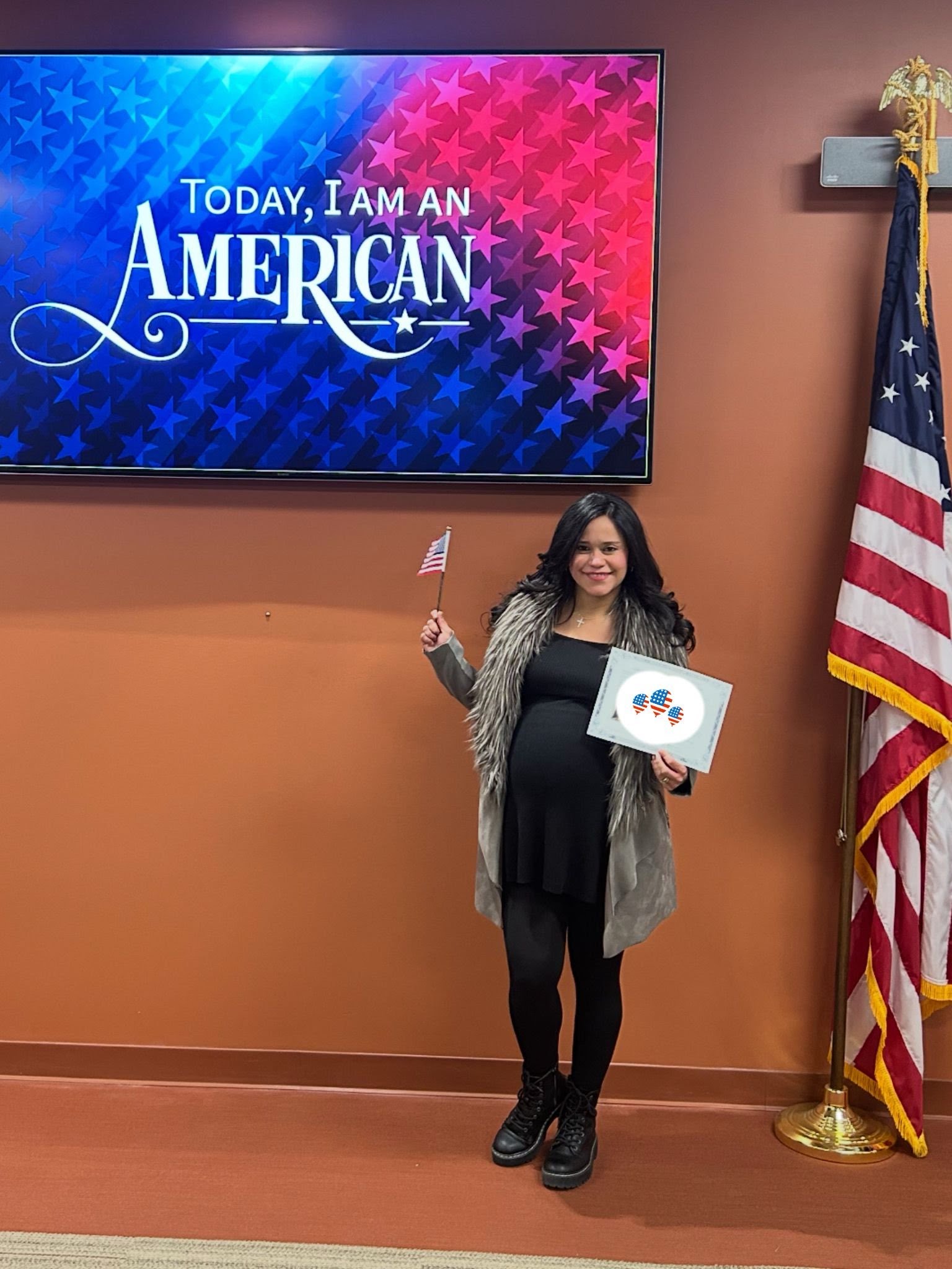 A woman stands stands next to a screen that says, "Today, I am an American" while holding an American flag.