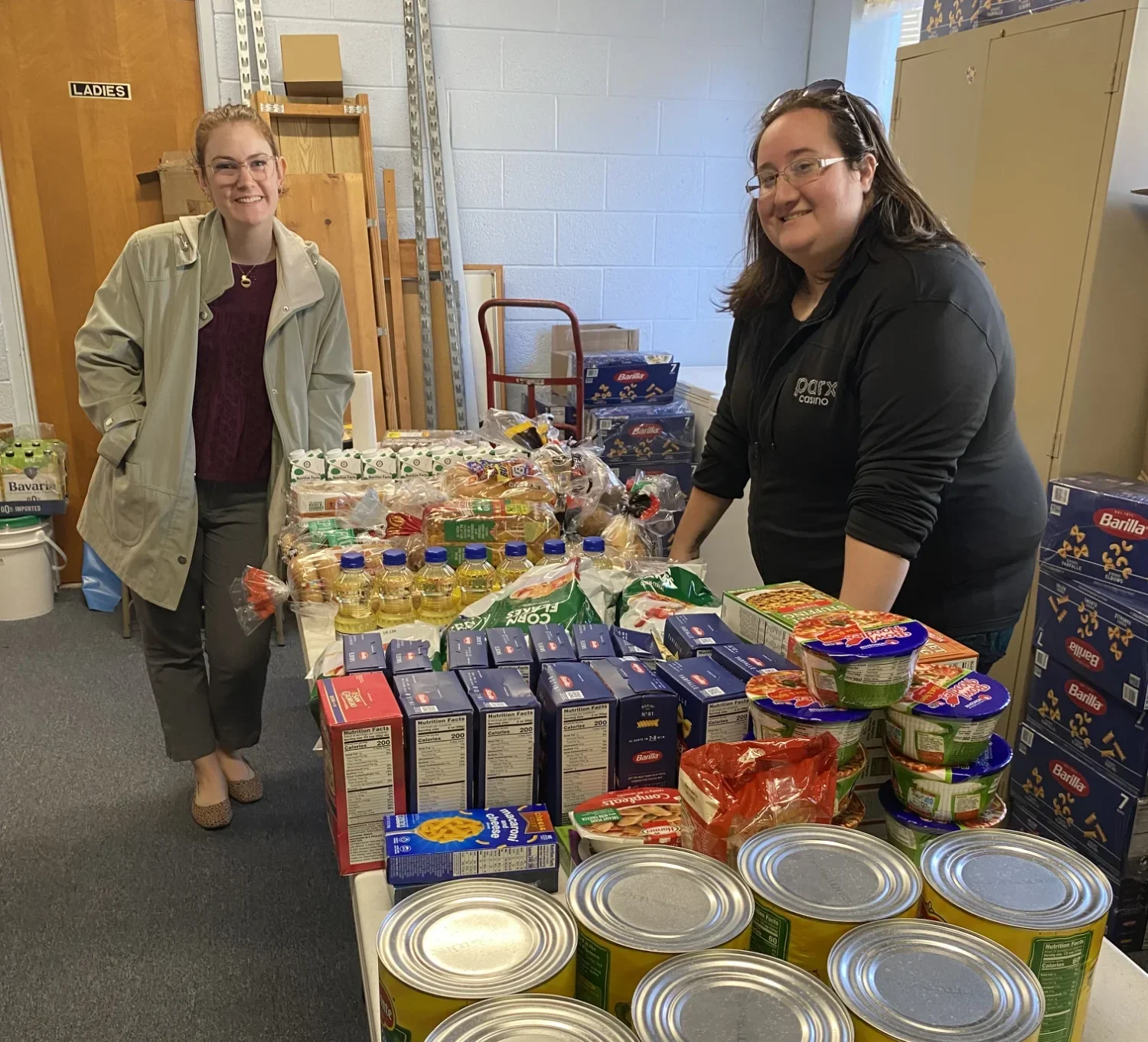 Two volunteers at a food pantry standing next to a table with food essentials.
