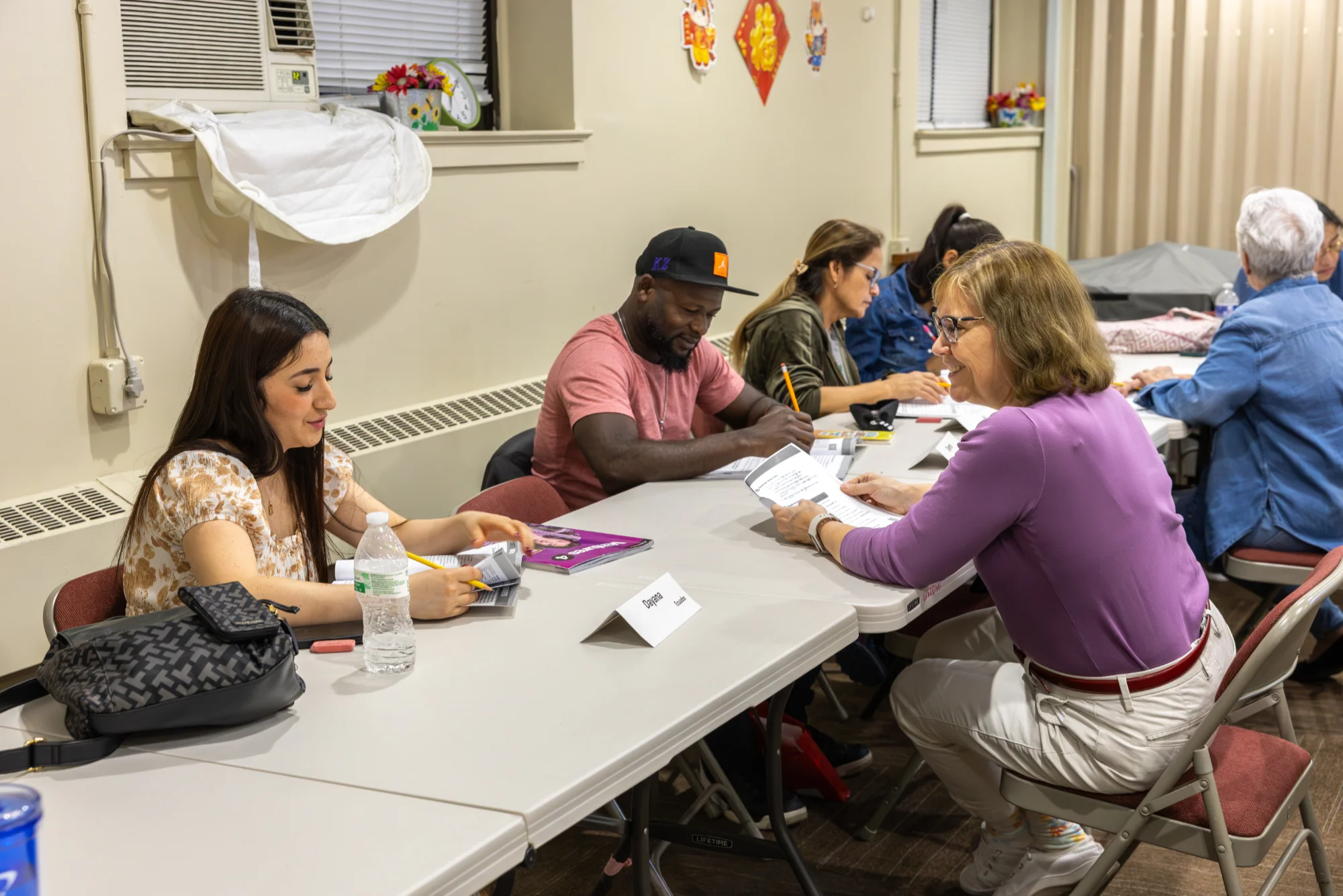 A group of students working through ESL exercises in their workbook as a teacher sits with them and answers their questions.