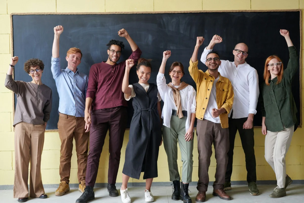 A group of students standing together in a classroom smiling and putting their hands in the air.
